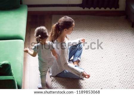 Young single mother doing yoga exercise, meditating while little preschool daughter running, playing, no stress, relax, tired mum sitting in yoga pose, emotions control, motivation, calm mind concept Royalty-Free Stock Photo #1189853068