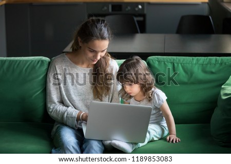 Single mother using laptop together with little preschool daughter, girl watching at computer screen, babysitter watching cartoons with pupil, showing training materials. Top above view
