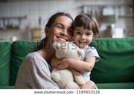 Smiling single young mum embracing little preschool daughter with toy, playing in living room at home, mother laughing with child, headshot portrait, cute girl look at camera Royalty-Free Stock Photo #1189853128