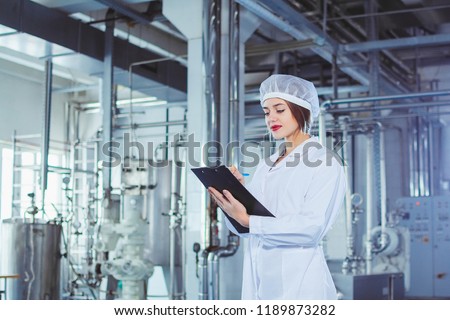 A young beautiful girl in white overalls makes notes in a tablet on the background of equipment of a food processing plant. Quality control in production. Royalty-Free Stock Photo #1189873282