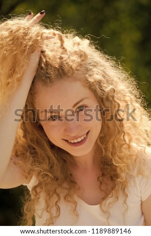 Portrait of one cute positive charming blonde young smiling woman with pleasant joyful face holding long curly hair with hand sunny day outdoor on natural background, vertical picture