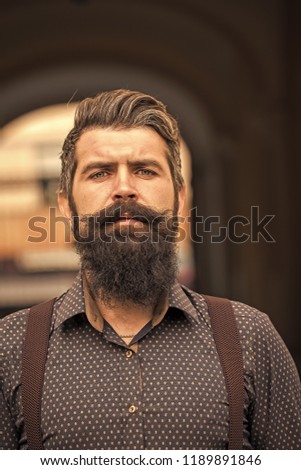Closeup view of one handsome senior stylish man with black hair and long lush beard in blue shirt standing outdoor on street arch background, vertical picture