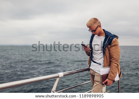 College student with backpack and book taking picture of sea using phone and admiring view on beach in Odessa