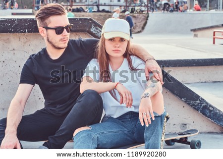 Trendy dressed couple of young skaters hugging together while sitting on skateboards at skatepark on sunny day.