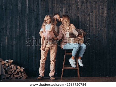 Family concept. Handsome father holds daughter on hands and kiss his wife with gifts in empty room against a wooden wall.