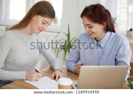 Two positive happy millennial young business women sitting at the desk in office and signing a contract. Client and broker agent, lease agreement, successful deal buy sell property real estate concept