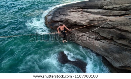 Aerial Photo of Mindfulness Scenery: a Boulder With Brazilian Atlantic Ocean Cliff and a Young Bravery Man Sitting on a Tightrope, Waterline Chalenge in a Secret Paradisiac Beach of Rio de Janeiro Royalty-Free Stock Photo #1189980100