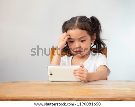 Little cute girl sitting on chair and use hand scratching a head. While she watching smartphone with seriously. If children watches a phone too much they may have ADHD.