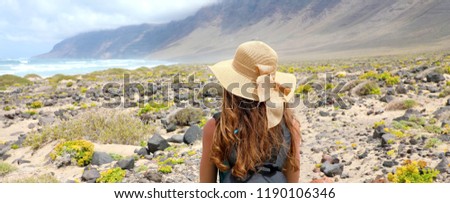 Rear view of traveler girl with straw hat looking at beautiful natural landscape. Young female backpacker exploring Lanzarote, Canary Islands. Banner panorama.