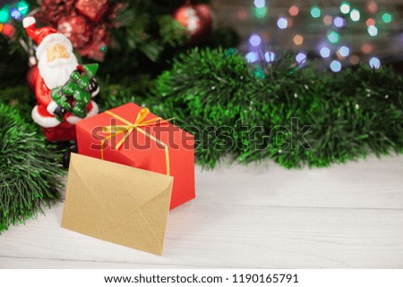 Christmas gift in red packing with a gold envelope next to a Santa Claus figure next to a Christmas tree and red Christmas balls with a ball on the background of a colored garland