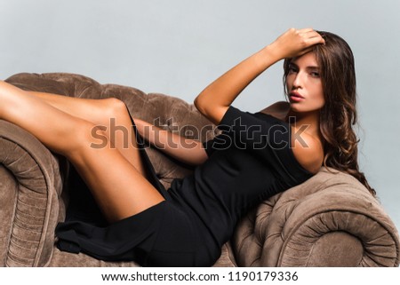 Gorgeous babe. Beautiful young woman in black dress looking at camera while sitting against white background in big chair
