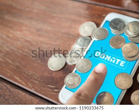 Finger pressing donate icon button on blue screen on white mobile phone with coins Thai baht money on wooden background with copy space. Donation online concept.