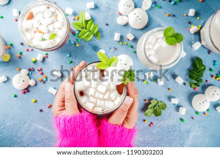 Girl drinks hot chocolate with funny marshmallow in form snowmen, white bears, with sweets and decorative leaves, light blue background, top view copy space, hands in picture