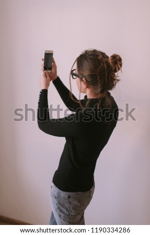 Making selfie photo, looking into camera, isolated. Girl with positive emotion. Young attractive brunette woman in black clothes wearing glasses holding smart phone, taking a photo. 