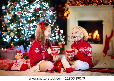 Child under Christmas tree at home. Little boy and girl in knitted sweater with Xmas ornament drink hot chocolate. Family with kids celebrate winter holidays. Kids open presents at fireplace.