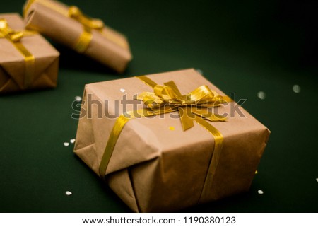Boxes with gifts for the holiday wrapped in Kraft paper with gold bows lie on a dark green background among confetti.