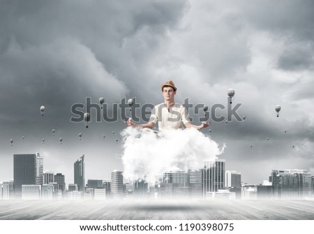 Man in white clothing keeping eyes closed and looking concentrated while meditating on cloud with cityscape view and flying aerostats on background.