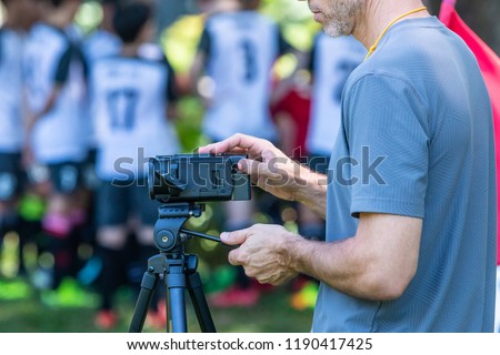 Man recording football match with his video camera on a tripod at an outdoor field with blurred players and trees background