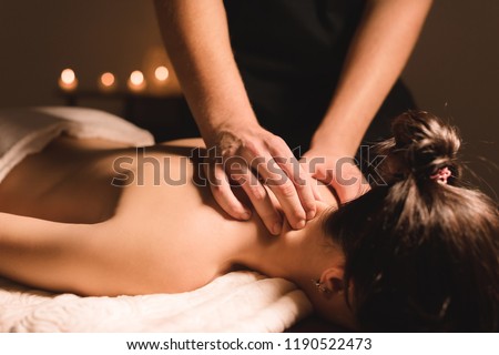 Men's hands make a therapeutic neck massage for a girl lying on a massage couch in a massage spa with dark lighting. Close-up. Dark Key Royalty-Free Stock Photo #1190522473