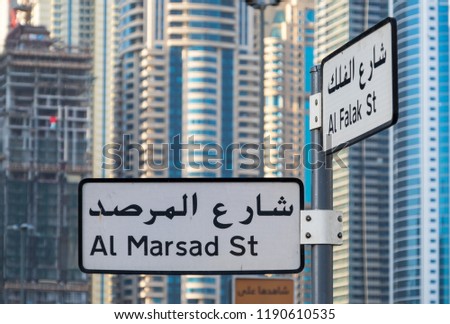Two street signs with Arabic writings in front of a crowded background made of tall skyscrapers