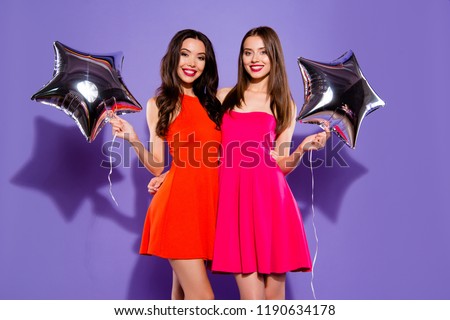 Portrait of two good-looking, magnificent, exquisite, delicate ladies with modern hairdro in fuchsia, red formal wear stand isolated on vivid violet background with stars helium blow decoration