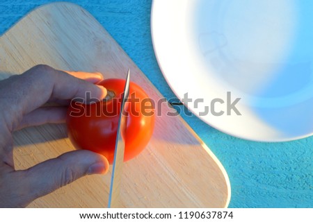 cutting a tomato on a cutting board on a blue-turquoise table near white ceramic plate