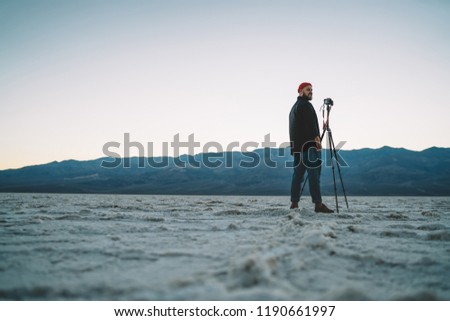 Young male photographer standing near tripod with camera waiting for sunset for shooting video, rear view hipster guy making pictures of wild nature and landscapes in desolate lands of death valley