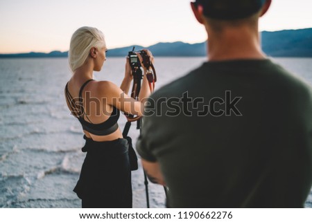 Young woman standing near tripod with equipment shooting video of nature environment standing near male colleague, back view of female travel photographer shooting video using camera  in journey
