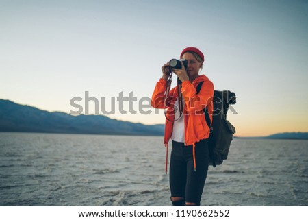 Tourist female photographer in bright orange jacket making picture of sunset sky in desert during journey, young woman taking photos in twilight on professional camera exploring nature on vacation