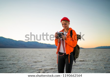 Smiling tourist hipster girl in orange jacket holding camera for viewing photos spending time in vacations, professional photographer using equipment for making pictures of wild environment