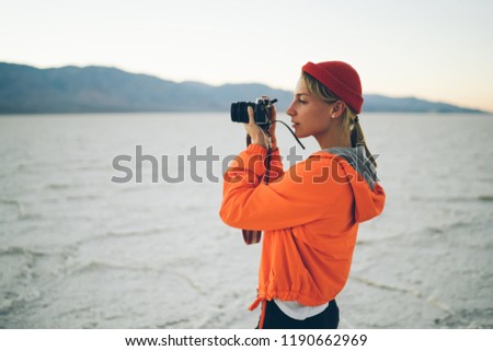 Female tourist fond of photography using modern equipment lens for taking pictures of nature and landscape in desert, young woman in orange jacket making photos during vacations travel expedition