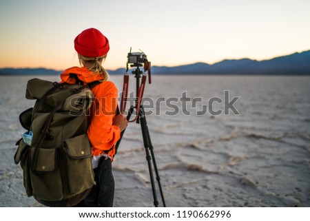 Hipster girl with touristic backpack using professional equipment for taking photos in evening desert, back view of young woman photographer standing near tripod with camera for make nature pictures