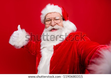 Aged mature stylish grandfather Santa Claus with white beard in costume headwear gloves stylish spectacles show like sign thumb up take picture on camera of smartphone isolated on red background