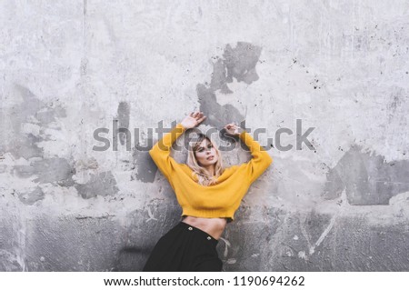 Blond girl in a yellow sweater and in a black skirt against a gray wall background