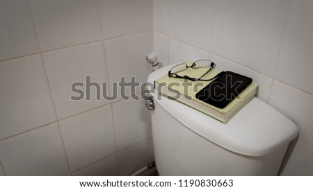 Black mobile phone Glasses and books are placed on the toilet bowl in the bathroom.