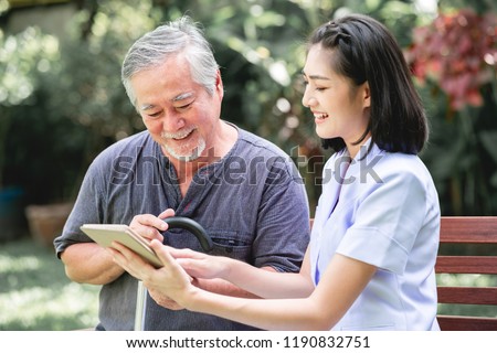 Nurse with patient sitting on bench together looking at tablet. Asian old man and young woman sitting together talking. Relax mood. Royalty-Free Stock Photo #1190832751