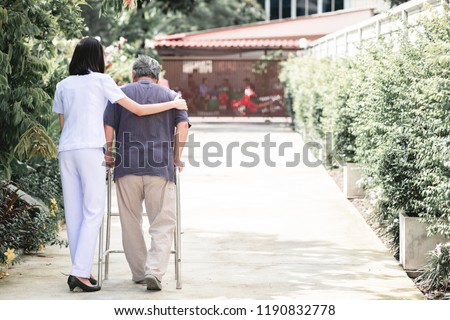 Nurse with patient using walker in retirement home. Young female nurse holding old man's shoulder in outdoor garden walking. Senior care, care taker and senior retirement home service concept. Royalty-Free Stock Photo #1190832778