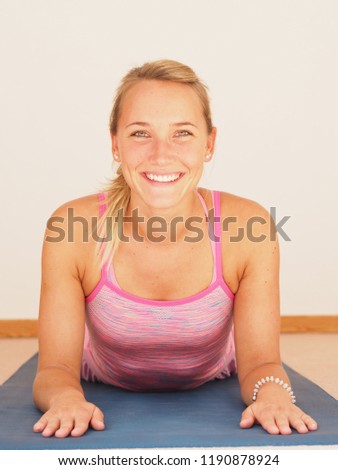Sporty young woman smilimg while doing sphinx pose, practicing yoga, baby cobra pose, working out, wearing sportswear, pink top, indoors, only upper body from front