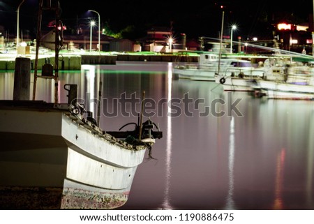 An old, small and white fishing boat parked at night on the shore. Long exposure shot and water is purplish color with reflection of lights. 