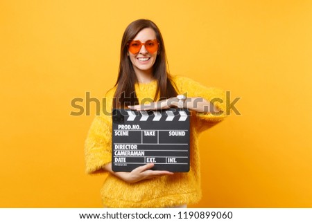 Portrait of smiling young woman in fur sweater, orange heart eyeglasses hold classic black film making clapperboard isolated on yellow background. People sincere emotions, lifestyle. Advertising area