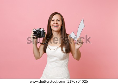 Smiling funny bride woman in wedding dress holding retro vintage photo camera and check mark, choosing staff, photographer isolated on pink background. Wedding to do list. Organization of celebration