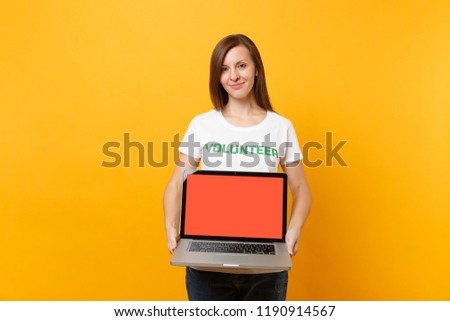 Woman in white t-shirt written inscription green title volunteer hold laptop pc computer, blank empty screen isolated on yellow background. Voluntary free assistance help, charity grace work concept