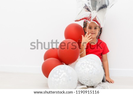 Happy brightful image of cute joyful little girl in tulle skirt sitting on present with balloons isolated on white background. Amazing charming birthday fashionable kid looking to camera