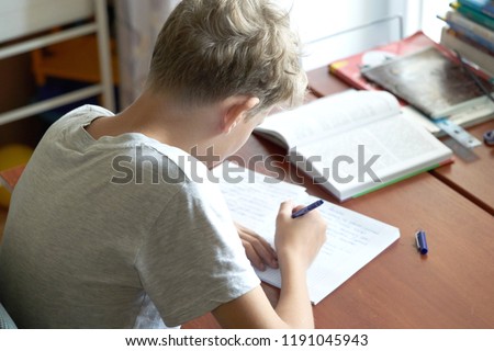 A cacausian blonde teenager boy with bad posture sitting at a table and writing in a copybook. Slouching blonde teenager boy doing school homework. Royalty-Free Stock Photo #1191045943