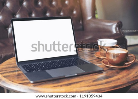 Mockup image of laptop with blank white desktop screen with coffee cups on wooden table in cafe