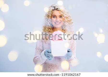 Picture showing curly blond woman in knitted sweater and gloves holding present box with pink bow indoor over gray wall background.