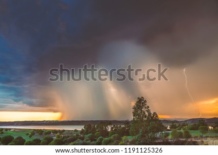 A massive rain cloud approaches as a storm rain breaks over Ayamonte, Andalusia Spain and the Guadiana river at sunset. The sky is golden behind the clouds and there is a lightning strike.