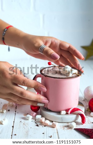 Female hands with Enamel cup of hot cocoa with mini marshmallows and candy canes with pine with beautiful Christmas toys. Christmas concept