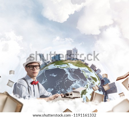 Side view of handsome man writer in hat and eyeglasses looking to camera while using typing machine at the table with flying books and Earth globe among cloudy skyscape on background. Elements of this