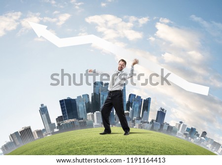 Young and confident businessman in suit starting launching huge white arrow to the air while standing on green lawn and cityscape view on background